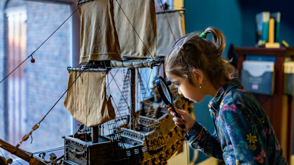 Meisje kijkt met vergrootglas naar modelschip 17e eeuw