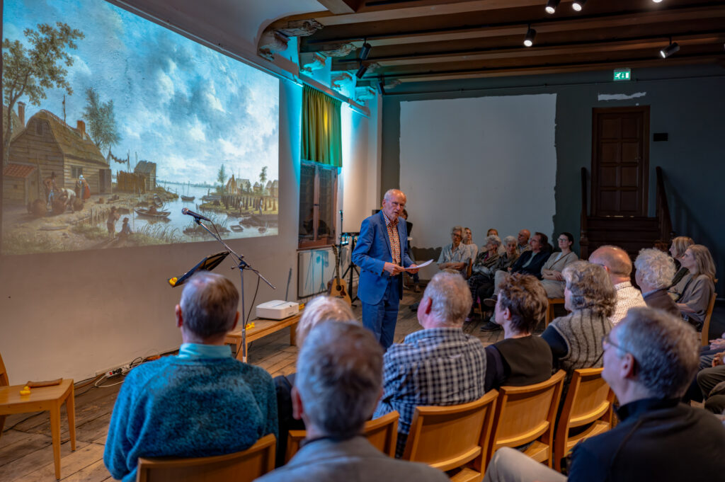 Persoon spreekt zaal met mensen toe met daarachter projectie van schilderij