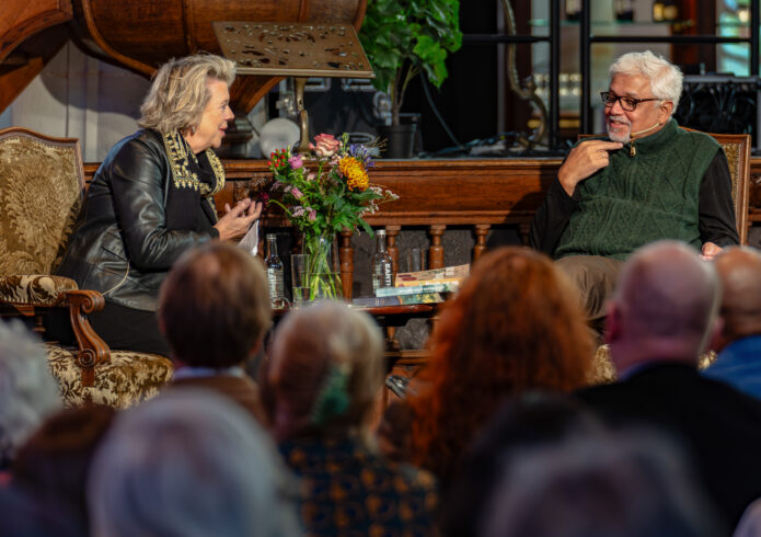 Twee personen in gesprek zittend in klassieke stoelen en ervoor publiek