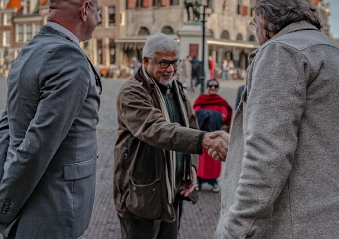 Drie personen waarvan twee de hand schudden op plein met oude gebouwen