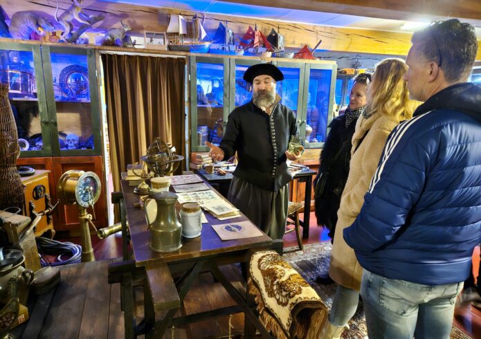 Persoon in 17e-eeuwse kleding geeft uitleg aan personen in oude kamer met balkenplafond
